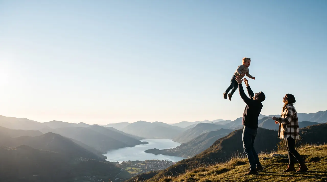 A parent lifts a laughing toddler into the air on a hilltop above a lake while another parent watches.